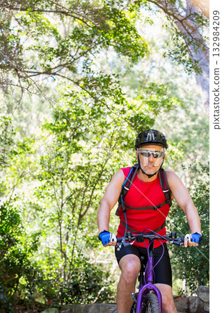 Mature adult man riding purple bike on forest trail wearing cycling helmet with hydration pack Mature adult man riding purple bike on forest trail wearing cycling helmet with hydration pack 132984209