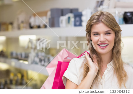 Woman holding pink shopping bags and smiling while browsing cosmetics shelves in beauty store aisle 132984212