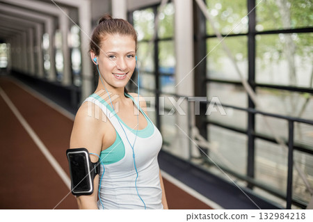 Female runner pausing on gym track lane, wearing tank top leggings phone armband and earphones 132984218