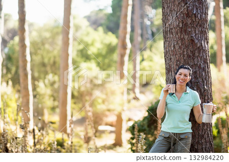 Woman leaning on pine trunk in forest clearing holding camping mug and coffee maker, copy space Woman leaning on pine trunk in forest clearing holding camping mug and coffee maker, copy space 132984220
