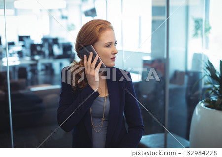 White businesswoman wearing navy blazer standing near glass partition in office, holding smartphone White businesswoman wearing navy blazer standing near glass partition in office, holding smartphone 132984228