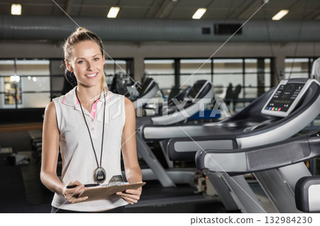 Female fitness trainer smiling beside treadmills in gym holding clipboard and pen wearing stopwatch Female fitness trainer smiling beside treadmills in gym holding clipboard and pen wearing stopwatch 132984230