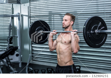 Shirtless male lifting barbell at shoulders in gym with cable machine and kettlebells, copy space 132984231