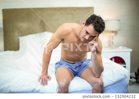 Shirtless man wearing blue shorts sitting on bed edge holding forehead by bedside lamp in bedroom Shirtless man wearing blue shorts sitting on bed edge holding forehead by bedside lamp in bedroom 132984232