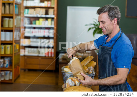 Male bakery clerk packaging loaves using tongs into bag at bakery counter wicker basket, copy space 132984234