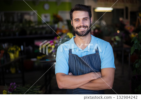 Man standing with crossed arms by wooden display tables in flower shop arranging colorful bouquets Man standing with crossed arms by wooden display tables in flower shop arranging colorful bouquets 132984242