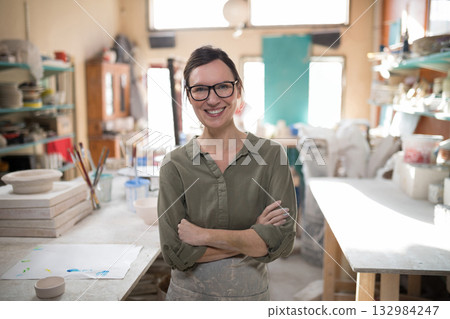Female potter wearing clay-stained apron standing in pottery studio near plaster molds, brush jars 132984247
