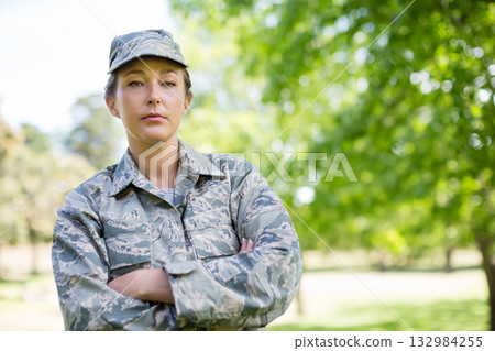 Mid adult female soldier standing with arms crossed in grassy park in camouflage uniform 132984255