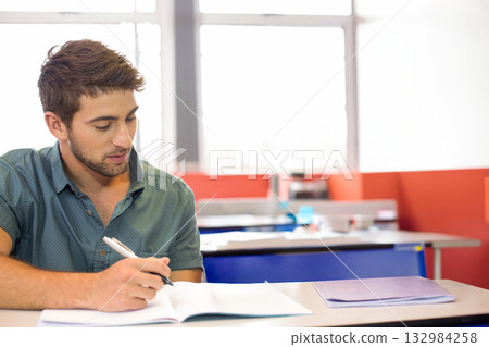 Male student sitting at classroom desk by window writing in open notebook with pen and folder 132984258