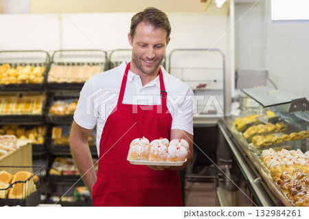 Male bakery worker wearing red apron standing at bakery counter holding wooden tray of pastries 132984261
