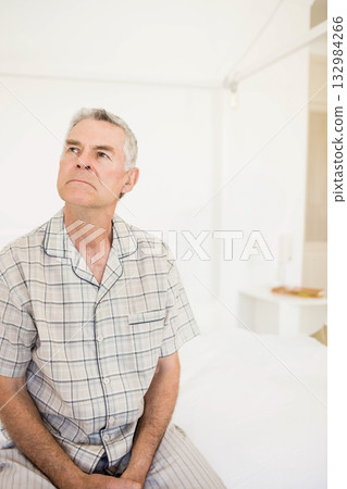 Senior man wearing plaid pajamas sitting on bed in bedroom beside table with lamp, copy space 132984266