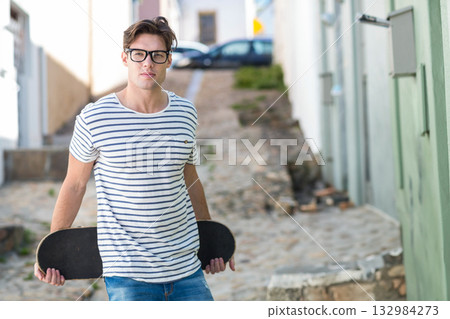 Teenage male holding skateboard behind back wearing eyeglasses on village sloped cobblestone lane 132984273