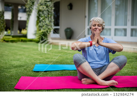 Senior woman practicing yoga on pink mat at veranda lawn with red fitness tracker, copy space Senior woman practicing yoga on pink mat at veranda lawn with red fitness tracker, copy space 132984274