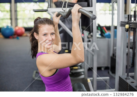 Woman performing lat pulldown exercise at gym in purple tank top using overhead cable machine 132984276