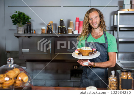 Man smiling, holding bundt cake on white stand wearing striped apron behind bakery counter 132984280