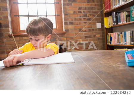 Boy sitting in study room at wooden table writing in spiral-bound notebook with pencil, copy space Boy sitting in study room at wooden table writing in spiral-bound notebook with pencil, copy space 132984283