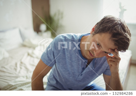 Man sitting on bed edge beside lamp and plant wearing blue henley shirt, copy space Man sitting on bed edge beside lamp and plant wearing blue henley shirt, copy space 132984286