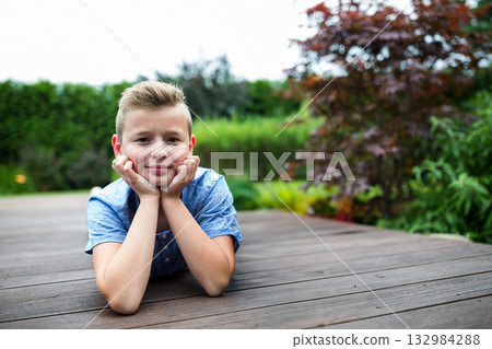 Boy lying on backyard deck resting chin on hands and gazing at camera with garden shrubs Boy lying on backyard deck resting chin on hands and gazing at camera with garden shrubs 132984288