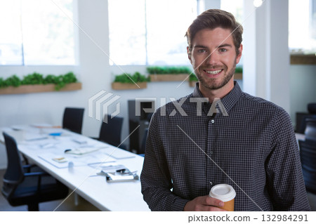 Man in thirties smiling while holding takeaway coffee cup at conference table in office, copy space Man in thirties smiling while holding takeaway coffee cup at conference table in office, copy space 132984291