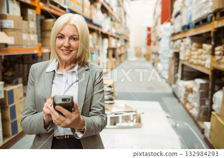 Woman in blazer scanning inventory with handheld scanner in warehouse aisle full of cardboard boxes 132984293