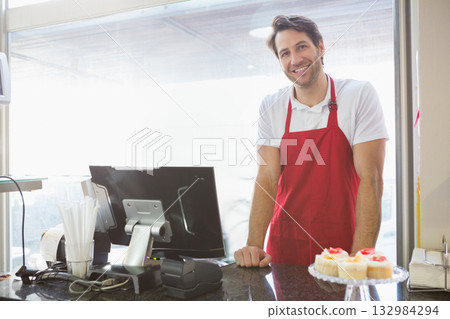 Mid adult male barista wearing red apron serving cupcakes at granite bakery counter, copy space 132984294