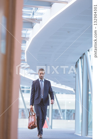 Businessman walking through curved office corridor in suit and carrying brown leather duffel bag Businessman walking through curved office corridor in suit and carrying brown leather duffel bag 132984301