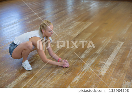 Woman squatting on wooden floor in dance studio wearing white sneakers, pink wristband, copy space Woman squatting on wooden floor in dance studio wearing white sneakers, pink wristband, copy space 132984308