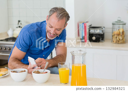 Man in mid-30s sitting at kitchen table checking smartphone while eating cereal and drinking juice Man in mid-30s sitting at kitchen table checking smartphone while eating cereal and drinking juice 132984321