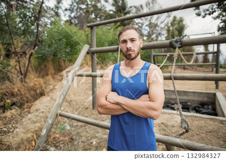 Fit man in sportswear on training field facing obstacle course with hanging ropes and tire pit 132984327