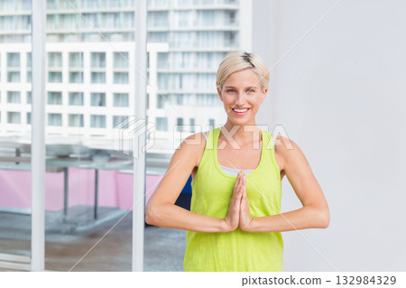 Smiling woman pressing hands together in fitness studio on grey floor by glass window 132984329