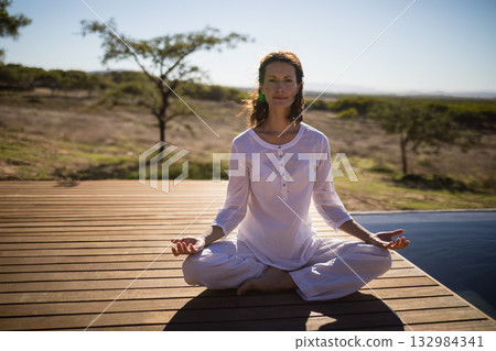 Woman sitting cross-legged on wooden deck by pool practicing meditation in white tunic and pants Woman sitting cross-legged on wooden deck by pool practicing meditation in white tunic and pants 132984341