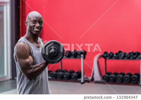 African American man performing bicep curl holding dumbbell at gym with rack, red wall, copy space African American man performing bicep curl holding dumbbell at gym with rack, red wall, copy space 132984347