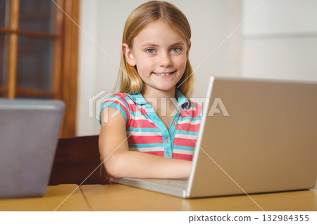 Child girl smiling directly at camera while sitting at wooden study table using silver laptop 132984355