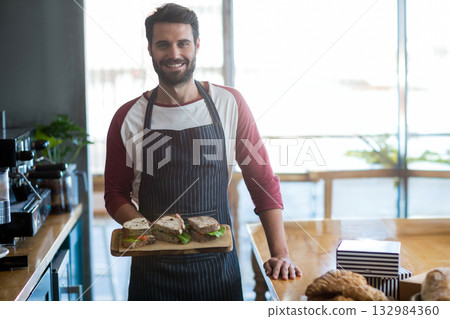 Male barista standing in cafe holding board of lettuce-topped sandwiches beside espresso machine Male barista standing in cafe holding board of lettuce-topped sandwiches beside espresso machine 132984360