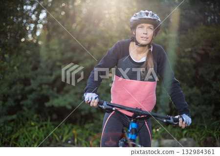 Woman cyclist riding mountain bike along wooded trail in sunlight wearing helmet, fingerless gloves Woman cyclist riding mountain bike along wooded trail in sunlight wearing helmet, fingerless gloves 132984361