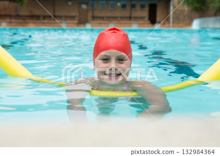 Girl wearing red swim cap and pink straps floating in outdoor pool holding yellow pool noodle Girl wearing red swim cap and pink straps floating in outdoor pool holding yellow pool noodle 132984364