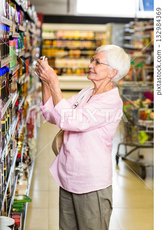 Senior female shopper reaching for jar on supermarket aisle shelves while pushing cart with produce Senior female shopper reaching for jar on supermarket aisle shelves while pushing cart with produce 132984369
