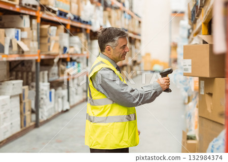 Male warehouse worker scanning cardboard boxes on metal shelving in aisle with handheld scanner 132984374