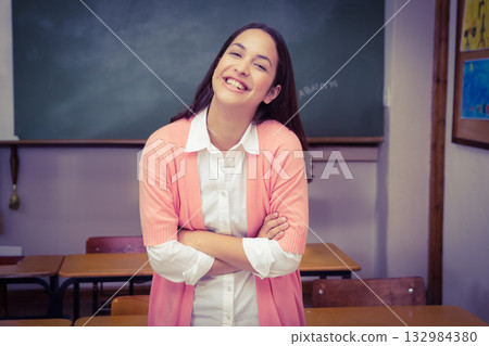 Female teacher standing in classroom crossing arms smiling at chalkboard, desks and bulletin board 132984380