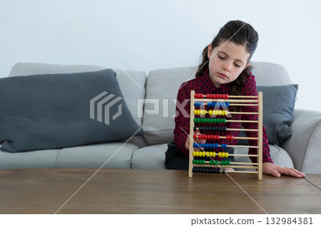 Female school-age child sitting on light grey sofa in living room using wooden abacus, copy space 132984381