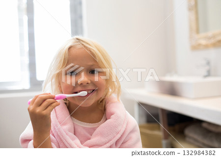 Female child brushing teeth at bathroom sink in pink bathrobe with pink toothbrush and gold mirror 132984382