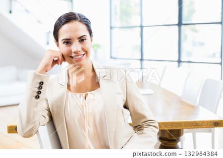 Female professional sitting at wooden table in office with pale sofa, staircase, smiling Female professional sitting at wooden table in office with pale sofa, staircase, smiling 132984384