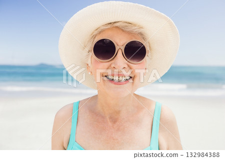 Senior woman smiling on white sandy beach wearing straw hat round sunglasses turquoise swimsuit 132984388