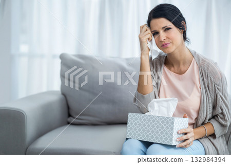 Woman sitting on grey sofa wiping tears with patterned tissue box in living room, copy space 132984394