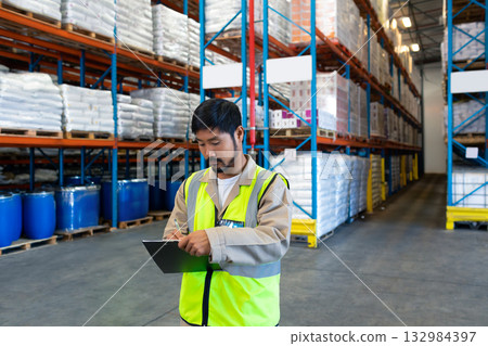 Asian man in his 30s wearing safety vest inspecting inventory with tablet among warehouse shelves 132984397
