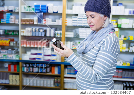 Mature adult woman holding dark medicine bottle in pharmacy aisle reading label, copy space 132984407