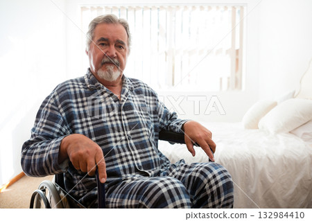 Senior man sitting in wheelchair wearing pajamas in bedroom by window blinds near bed pillows 132984410