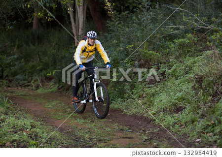 Male cyclist pedaling mossy forest trail wearing yellow jacket with mountain bike and red backpack Male cyclist pedaling mossy forest trail wearing yellow jacket with mountain bike and red backpack 132984419