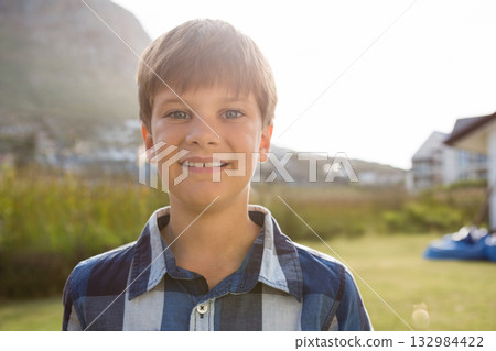 Preteen boy standing in grassy suburban field wearing blue and white plaid shirt under bright sun 132984422