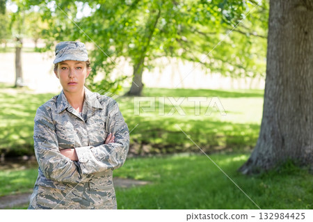 Mid adult female soldier standing with arms crossed in clearing wearing camouflage jacket and cap Mid adult female soldier standing with arms crossed in clearing wearing camouflage jacket and cap 132984425